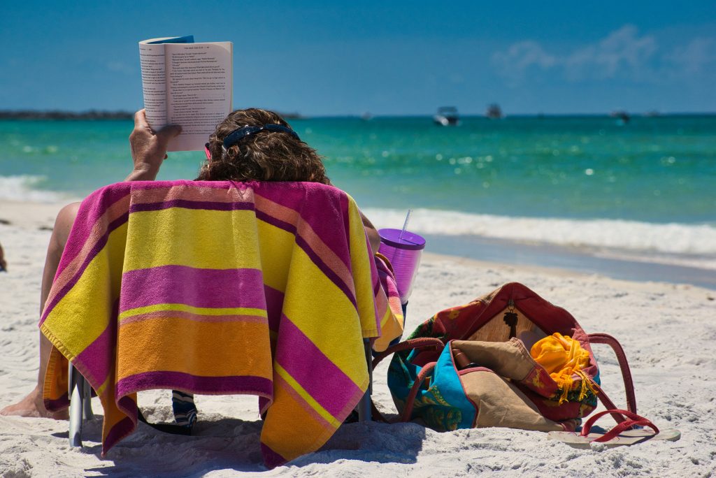 A person sits in a beach chair covered with a colorful striped towel, reading a book on a sunny sandy beach. A purple cup and a bag with belongings are beside them, with turquoise sea and boats in the background.